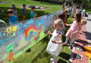 Dzieci w ogrodzie malują na folii, na obrazie widać słonaczko, błękitne chmurki, barwną tęczę oraz zieloną roślinność